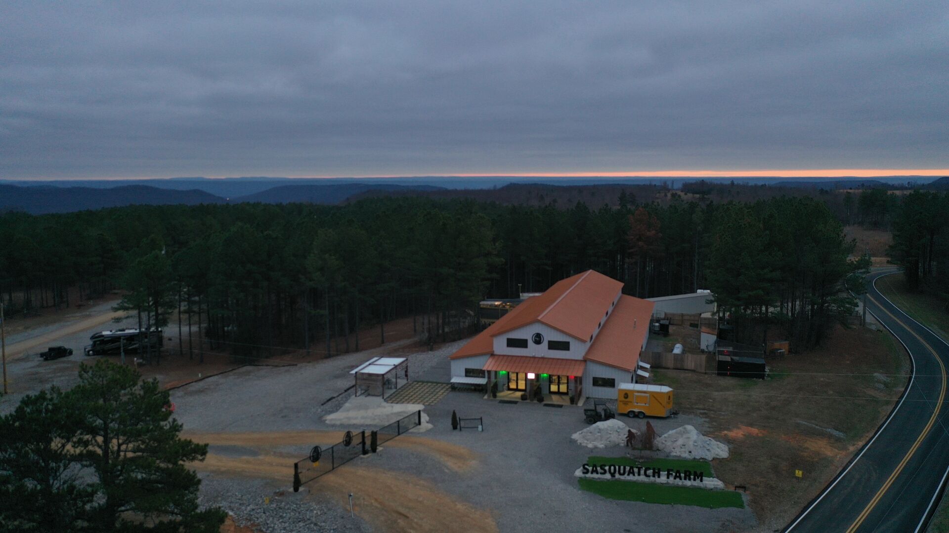 Aerial view of the Sasquatch Farm RV Park entrance gate and check-in building at dusk, with the road leading in